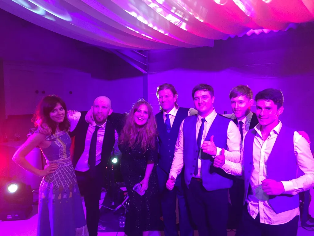 Group of seven people, dressed in formal attire, posing and smiling under purple and pink party lights.