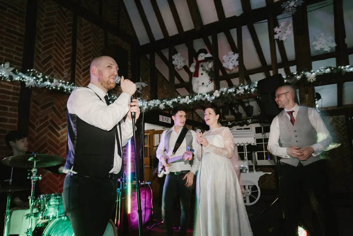 A band performing indoors decorated with garlands and snowflakes, featuring a man singing into a microphone, a woman in a white dress holding a microphone, a man playing bass guitar, a drummer, and another man smiling.