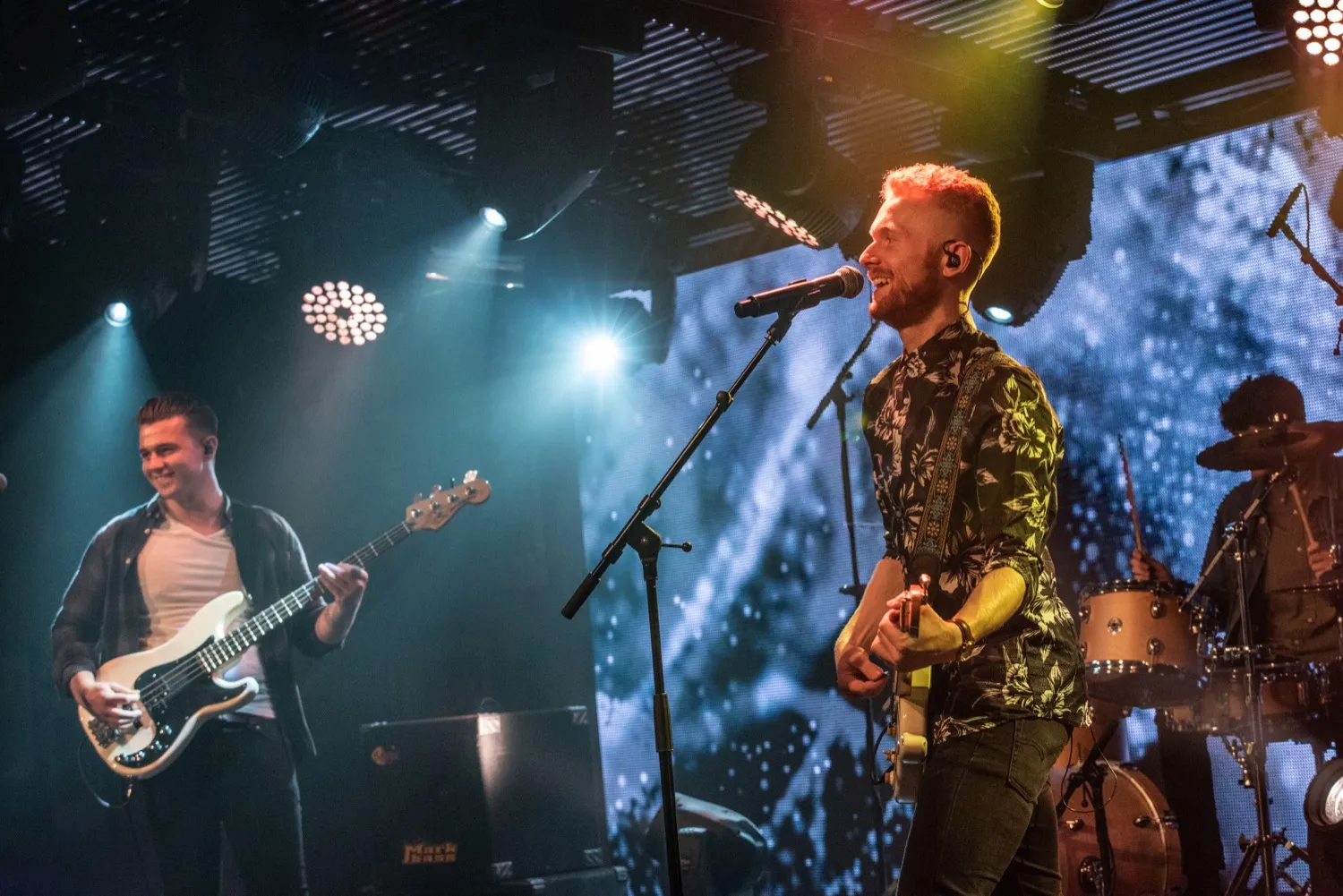 Three musicians performing on stage with guitars and drums under colorful stage lights.