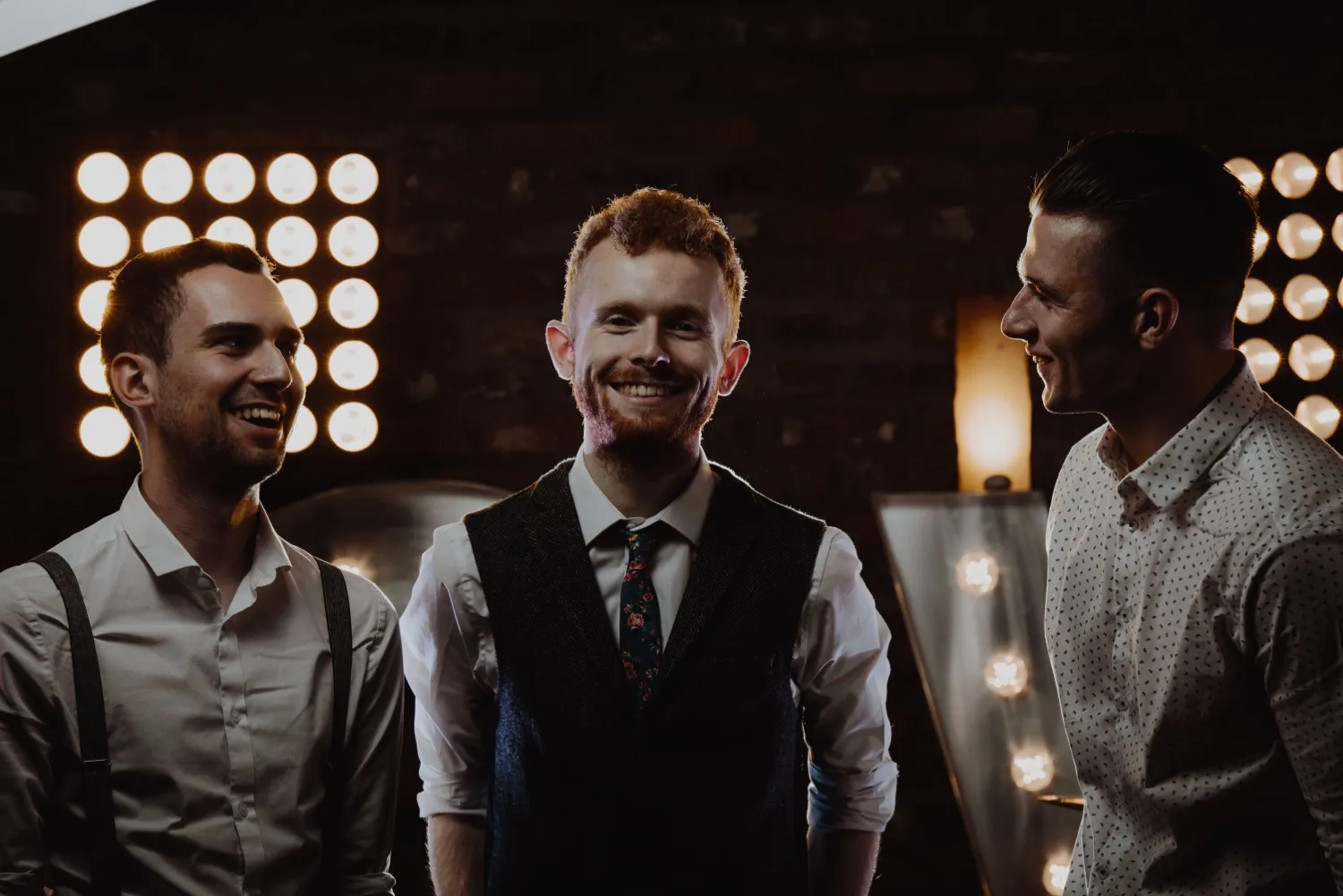 Three young men smiling and talking in a dimly lit room with round studio lights in the background.