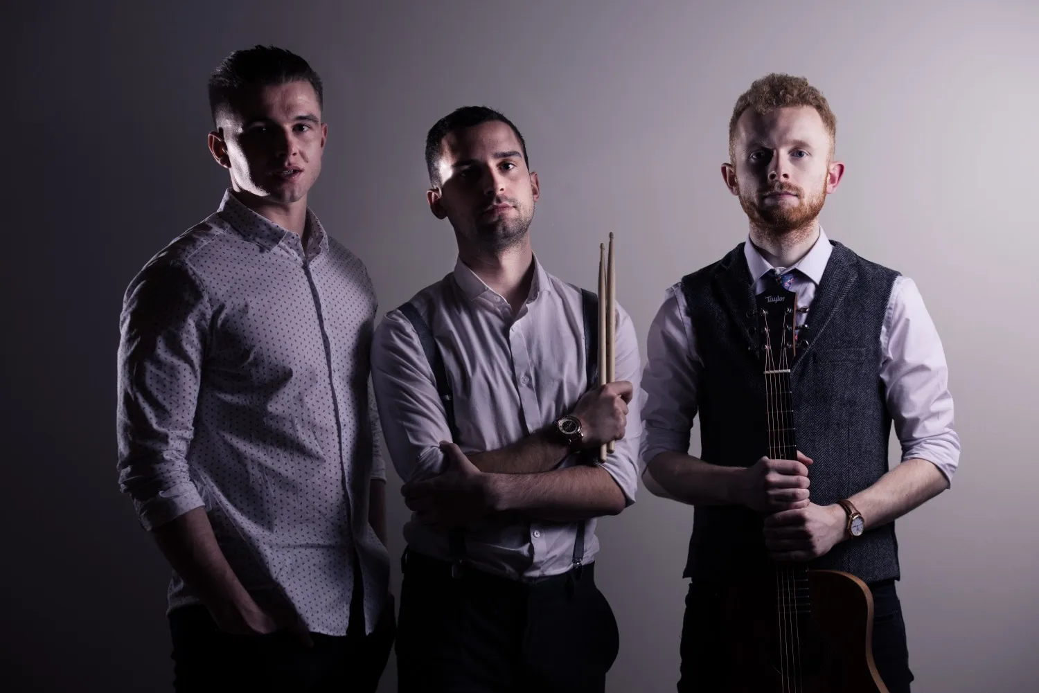 Three men posing against a gradient background; the left man wears a patterned shirt, the center man holds drumsticks, and the right man holds a guitar.