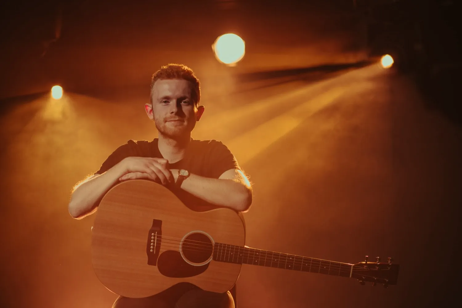 Young man sitting and smiling with his arms resting on an acoustic guitar under warm stage lighting.