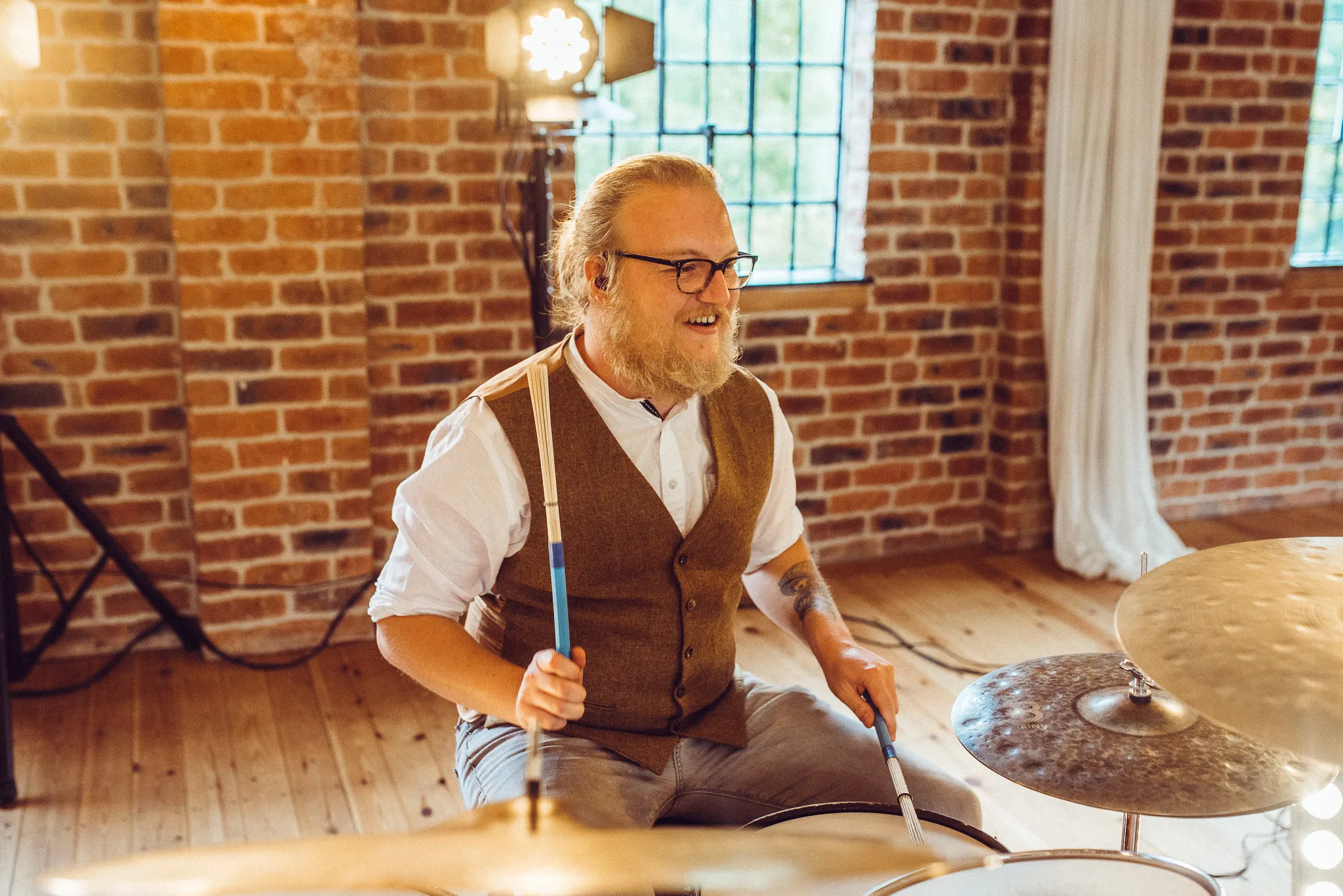 Bearded man with glasses and a vest playing drums in a room with brick walls and wooden floor.
